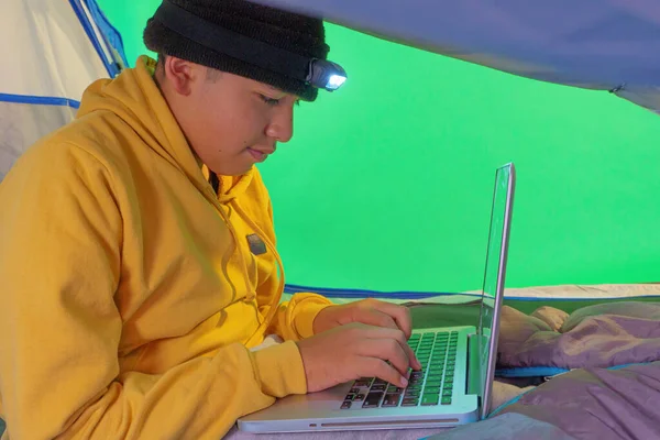 a boy playing with a laptop inside a tent on a green background