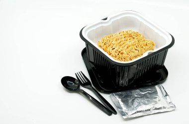 Siyah plastik bir kasede siyah plastik bir kaşık ve çatalla anlık erişteler, küçük bir folie paketindeki malzemeler, normal sıcaklıkta erişte çorbası..