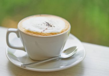 Sıcak cappuccino kahveyi doğal ışıkla kapat, ağacın yeşil renginin bulanık arkaplanı, seçilmiş odak. Küçük kaşık fincanın yanında..