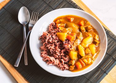 Yuvarlak beyaz tabakta pirinç meyveli Japon köri, doğal ışık ile sağlıklı yiyecekler, Japon köri ve kaşıkla pilav ve çatal siyah ahşap kaplama, yukarıdan görüş..