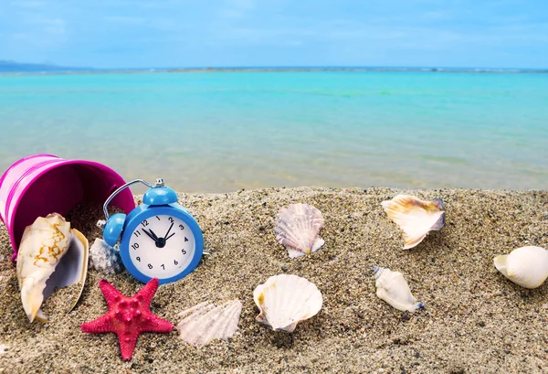 Conchas, relógio e balde na praia de areia com mar no fundo — Fotografia de Stock