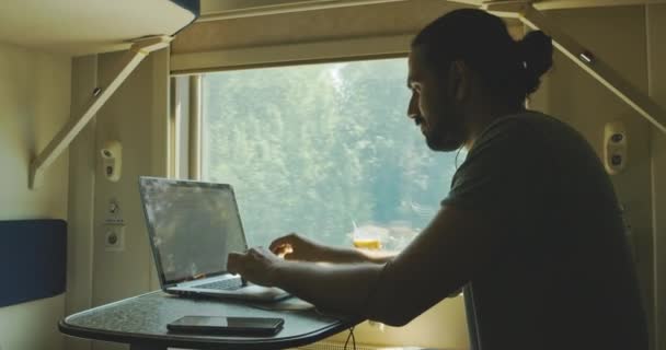 Un joven caucásico se sienta en auriculares con una computadora portátil mientras viaja en tren. — Vídeo de stock