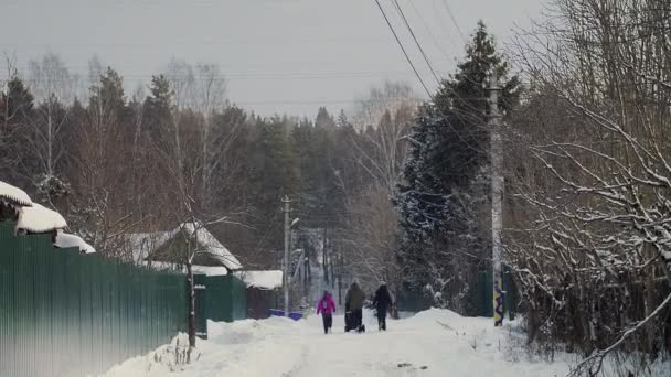 Снежная дорога в маленьком городке рядом с лесом. Люди и дети идут пешком — стоковое видео