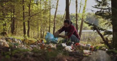 Erkek bir gönüllü, ormana saçılmış çöpleri plastik bir torbayla topluyor..