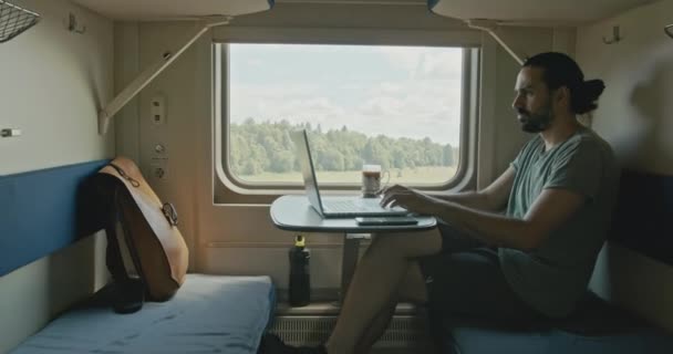 Un joven caucásico se sienta en auriculares con una computadora portátil mientras viaja en tren. Video de stock