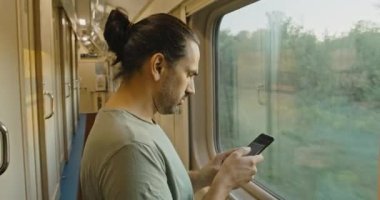Akıllı telefonlu genç bir turist hareket halindeki bir trenin camından dışarı bakar.