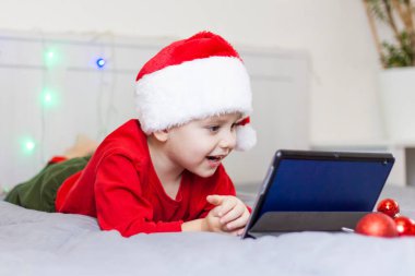 Tatlı bir çocuk yatakta oturuyor ve bir tablete bakıyor. Gülen komik çocuk. Noel Baba şapkası takıyordu. Noel