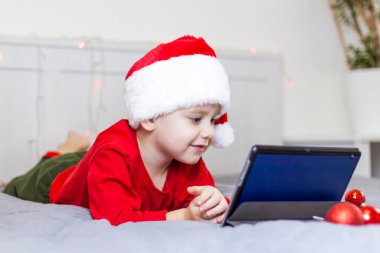 Tatlı bir çocuk yatakta tablete bakıyor. Gülen komik çocuk. Noel Baba şapkası takıyordu. Noel