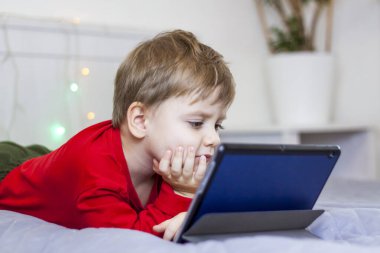 Yatakta tatlı bir çocuk tablete bakıyor. Çevrimiçi eğitim, internette iletişim. Gülen komik çocuk. Alet ve çocuk kavramı.