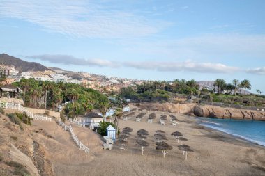 Playa del Duque, Costa Adeje, Tenerife, Kanarya Adaları, İspanya - 9 Ekim 2020: Öğleden sonra saatlerinde boş plaj, yakınlardaki kalabalık tatil beldeleri ve güneşliklerle çevrili.