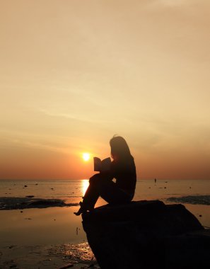 gün batımında sahilde kayada siluet kız okuma kitabı