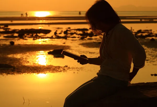 Mulher silhueta ler livro por praia ao nascer do sol — Fotografia de Stock