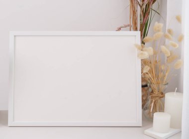 Kurutulmuş poster çerçevesi, modern cam vazoda Lagurus ovatus çiçekleri ve beyaz masa ve çimento duvar arka planında mumlar.