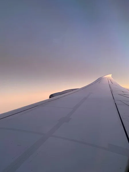 A colorful sunrise seen from the plane window. Sun is raising from the clouds, painting them pink and orange. Horizon line turns yellow with the sunbeams. Plane wing covering the sun.