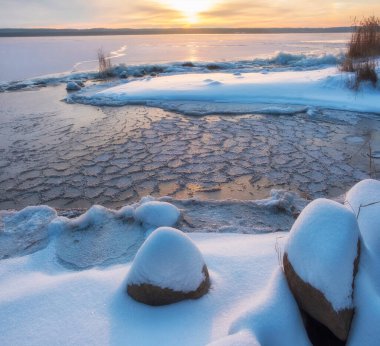 Kıyıdaki taze karın altında taşlar, kışın gün batımında buz ve karla kaplı göl.