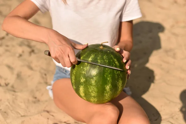 女の子はナイフで手に熟した緑色のスイカを持って切ります ビーチでピクニック 夏休みに長い脚をした若い女性が砂の上に座り — ストック写真