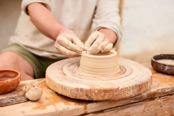 male craftsman master in light linen shirt sculpts jug, bowl or vase from clay in village