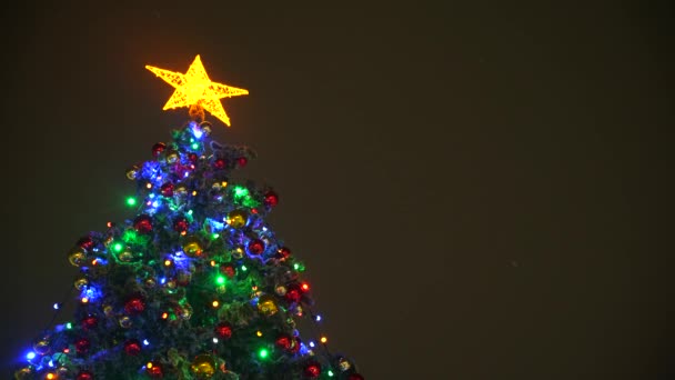 Árvore de Natal com uma estrela amarela, balões, brinquedos e luzes coloridas em um fundo escuro em uma queda de neve — Vídeo de Stock