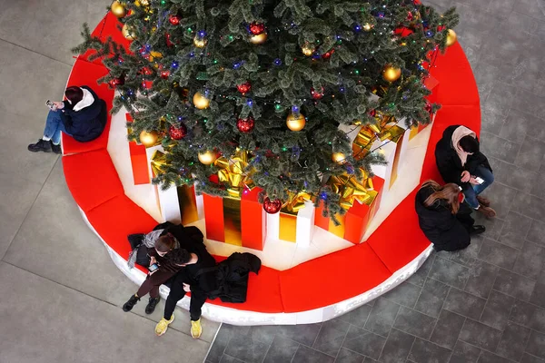 Young people are sitting on a bench near a Christmas tree decorated with garlands, lights, toys, yellow and red balloons