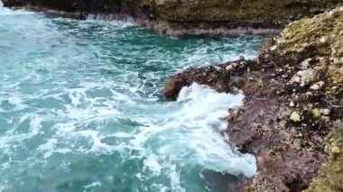 Deniz dalgaları deniz kıyısındaki taşlara çarpıyor. Mavi deniz, hafif fırtına, beyaz deniz köpüğü, güneşli hava.