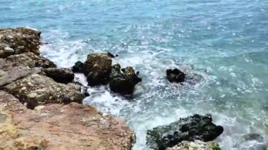 Deniz dalgaları deniz kıyısındaki taşlara çarpıyor. Mavi deniz, hafif fırtına, beyaz deniz köpüğü, güneşli hava.