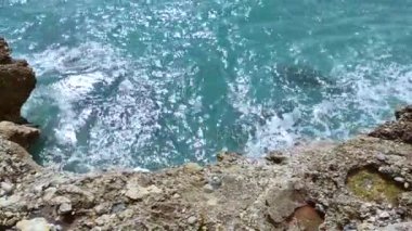 Deniz dalgaları deniz kıyısındaki taşlara çarpıyor. Mavi deniz, hafif fırtına, beyaz deniz köpüğü, güneşli hava.