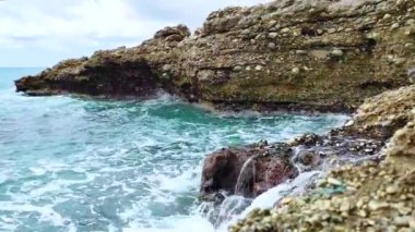 Deniz dalgaları deniz kıyısındaki taşlara çarpıyor. Mavi deniz, hafif fırtına, beyaz deniz köpüğü, güneşli hava.