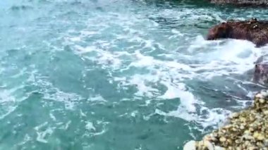 Deniz dalgaları deniz kıyısındaki taşlara çarpıyor. Mavi deniz, hafif fırtına, beyaz deniz köpüğü, güneşli hava.