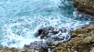 Deniz dalgaları deniz kıyısındaki taşlara çarpıyor. Mavi deniz, hafif fırtına, beyaz deniz köpüğü, güneşli hava.