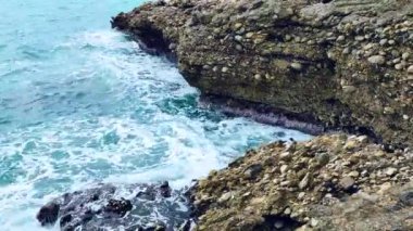 Deniz dalgaları deniz kıyısındaki taşlara çarpıyor. Mavi deniz, hafif fırtına, beyaz deniz köpüğü, güneşli hava.