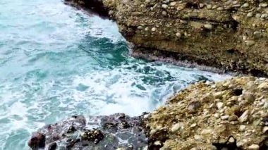 Deniz dalgaları deniz kıyısındaki taşlara çarpıyor. Mavi deniz, hafif fırtına, beyaz deniz köpüğü, güneşli hava.