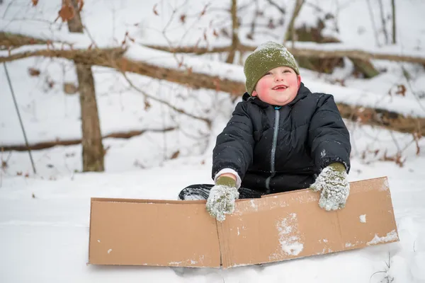 Niño jugando en la nieve usando caja de cartón para deslizarse colina abajo —  Fotos de Stock