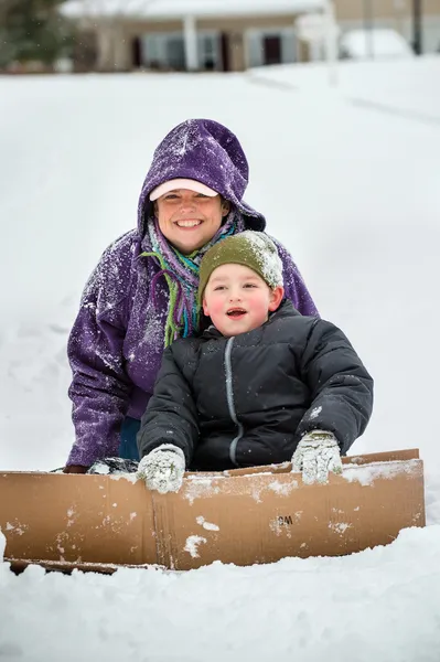 Mãe e filho brincando na neve usando caixa de papelão para deslizar colina abaixo — Fotografia de Stock