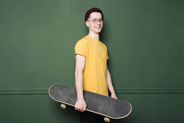 Fehér férfi sárga pólóban pózol korcsolyával át sötét zöld háttér fal — Stock Fotó
