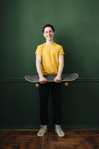 Blanke man in geel t shirt poseren met skate over donkergroene achtergrond muur — Stockfoto