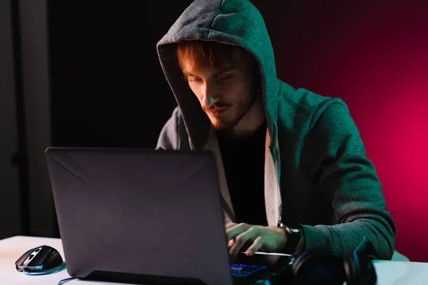 Professional hacker with laptop sitting at table on dark background — Stock Photo, Image
