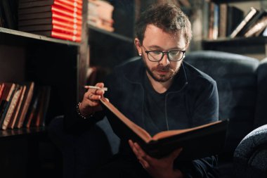 Yetişkin öğrenci üniversite kütüphanesinde sigara içerken kitap okuyor.