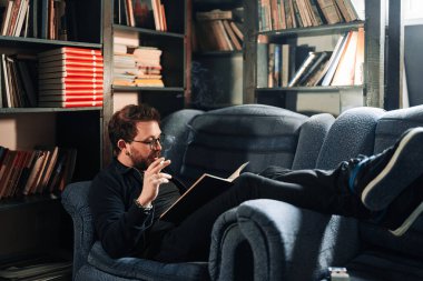 Yetişkin öğrenci üniversite kütüphanesinde sigara içerken kitap okuyor.