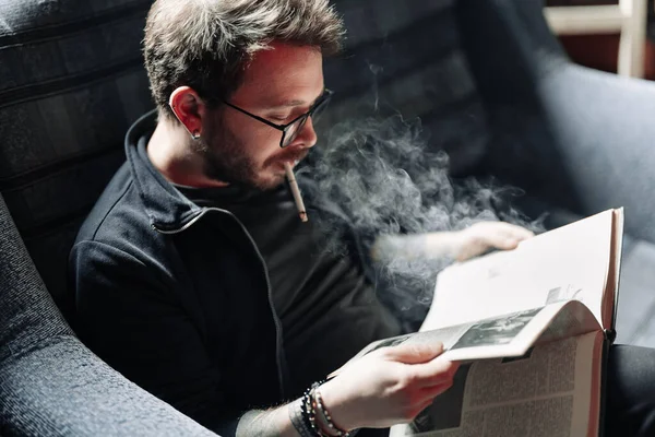 Estudiante adulto leyendo un libro en la biblioteca de la universidad mientras fuma — Foto de Stock