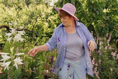 Güzel gülümseyen son sınıf kadını güneşli bir günde bahçede çiçek ve ağaçlardan hoşlanır.