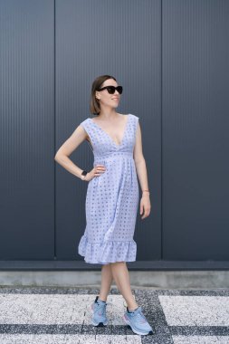 Young Caucasian Woman posing in blue dress on gray wall background outdoors. Female wearing sunglasses
