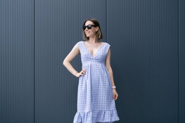 Young Caucasian Woman posing in blue dress on gray wall background outdoors. Female wearing sunglasses