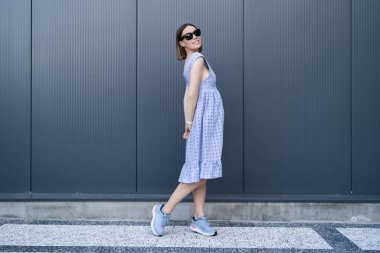 Young Caucasian Woman posing in blue dress on gray wall background outdoors. Female wearing sunglasses