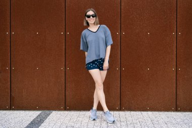 Young woman in casual t-shirt, shorts in sunglasses posing on rusty wall background. Urban style