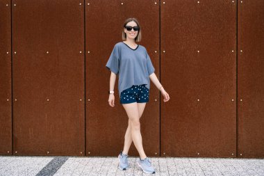 Young woman in casual t-shirt, shorts in sunglasses posing on rusty wall background. Urban style