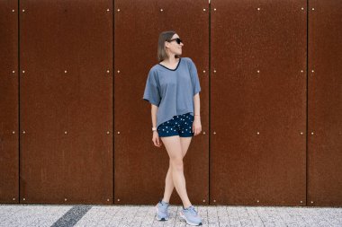 Young woman in casual t-shirt, shorts in sunglasses posing on rusty wall background. Urban style