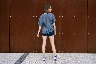 Young woman in casual t-shirt, shorts in sunglasses posing on rusty wall background. Urban style