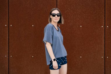 Young woman in casual t-shirt, shorts in sunglasses posing on rusty wall background. Urban style