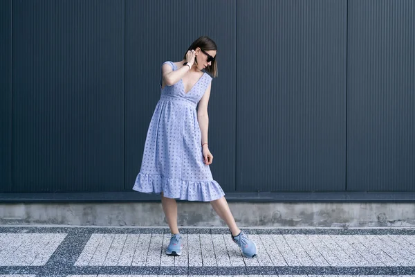 Young Caucasian Woman posing in blue dress on gray wall background outdoors. Female wearing sunglasses