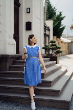 Mavi elbiseli güzel hamile bir kadın şehir sokaklarında yürüyor ve içki içiyor..